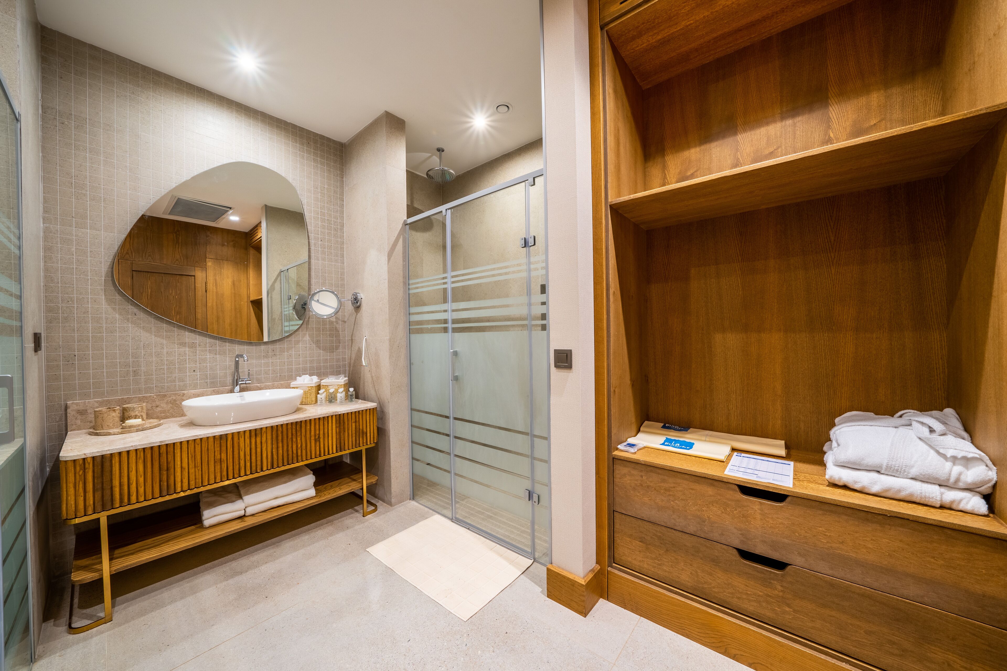 Un baño moderno con tocador de madera, un espejo grande, cabina de ducha de vidrio y un estante de madera con toallas.