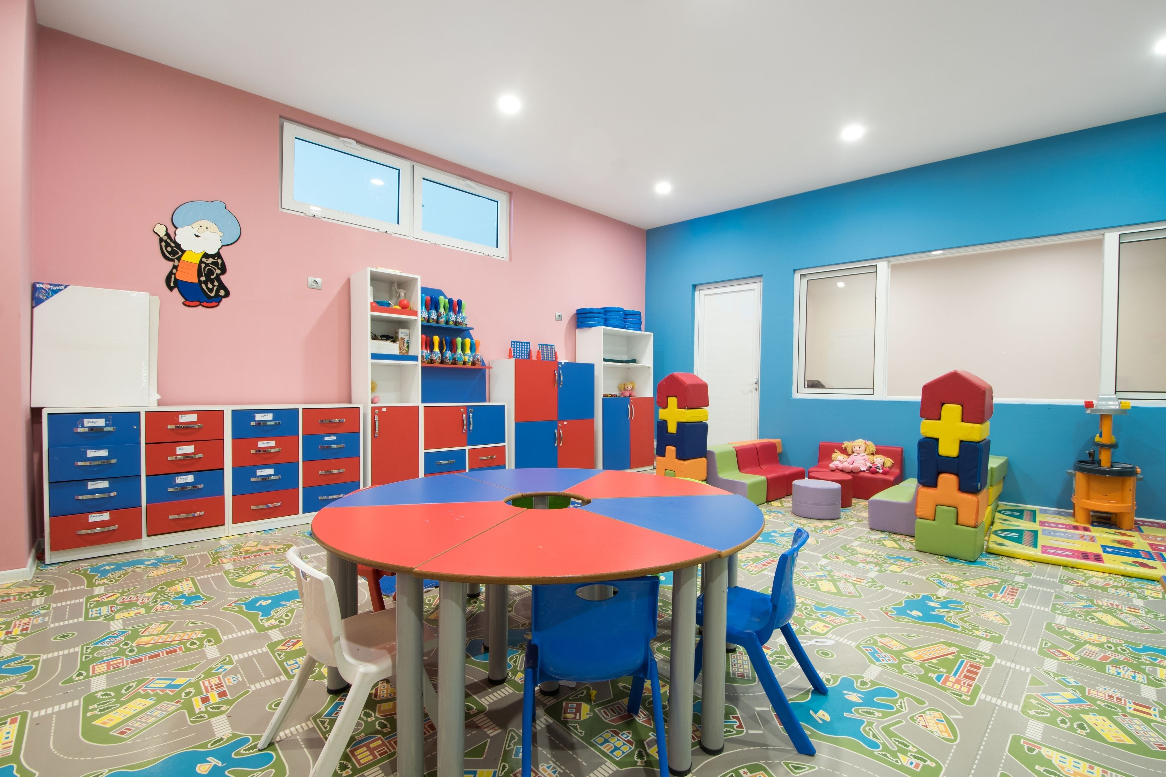 Un cuarto de juegos lleno de color, con una mesa redonda, sillas, armarios de almacenamiento y una zona de juego con bloques de construcción.