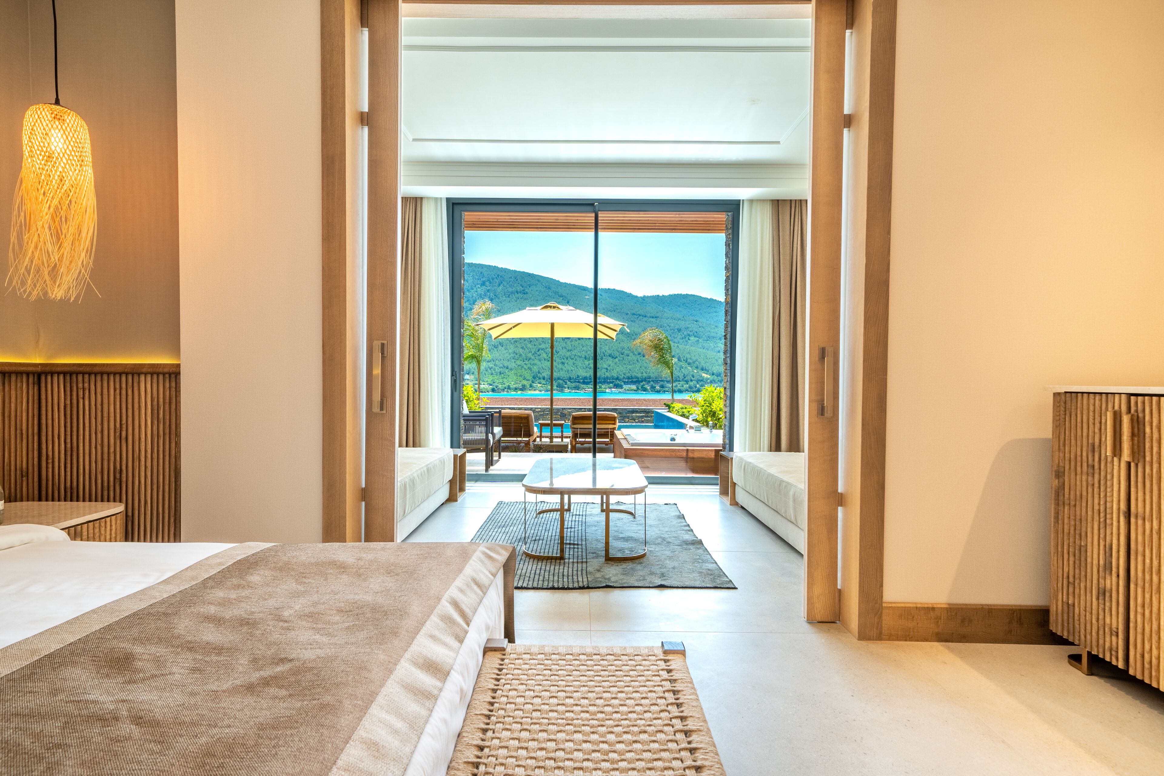 A modern hotel room with a large bed, a dining area with a table and chairs, and a view of a pool and mountains through large windows.