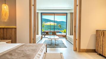 A modern hotel room with a large bed, a dining area with a table and chairs, and a view of a pool and mountains through large windows.