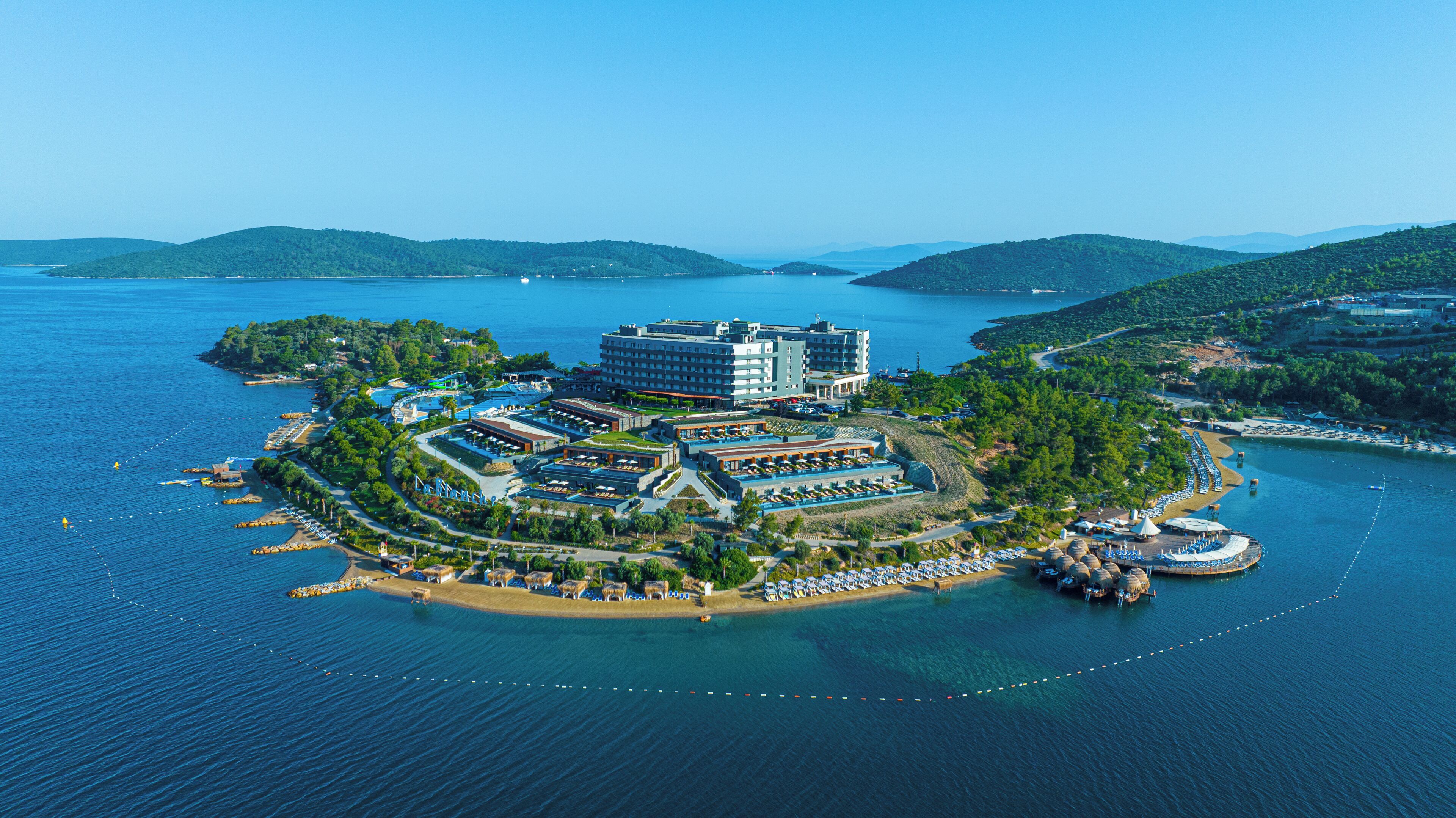 An aerial view of a coastal hotel with multiple buildings, a beach area with sun loungers, and a marina.