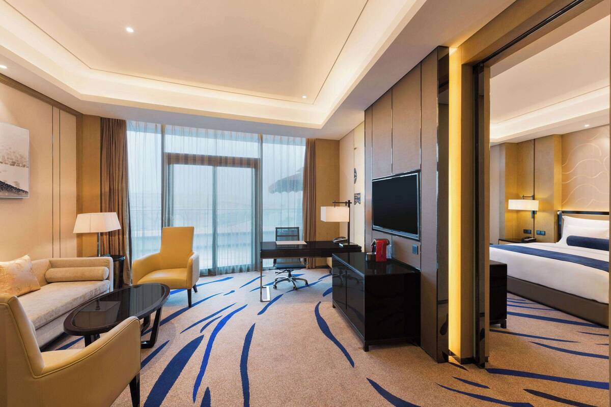 Una habitación de hotel con sofá, sillón, escritorio, televisión, cama y un ventanal grande.