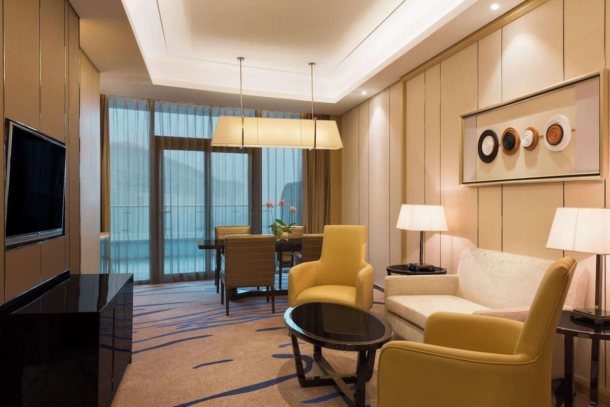 Habitación de hotel con televisor de pantalla plana, sofá, dos sillones, una mesa de comedor con sillas y un ventanal con cortinas.