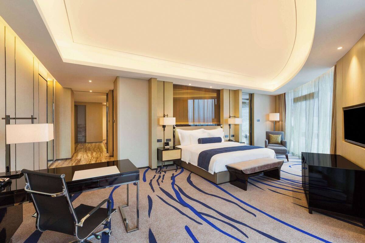 Una habitación de hotel moderna con una cama grande, un escritorio, una silla, un televisor y un baño visible a través de una puerta de vidrio.