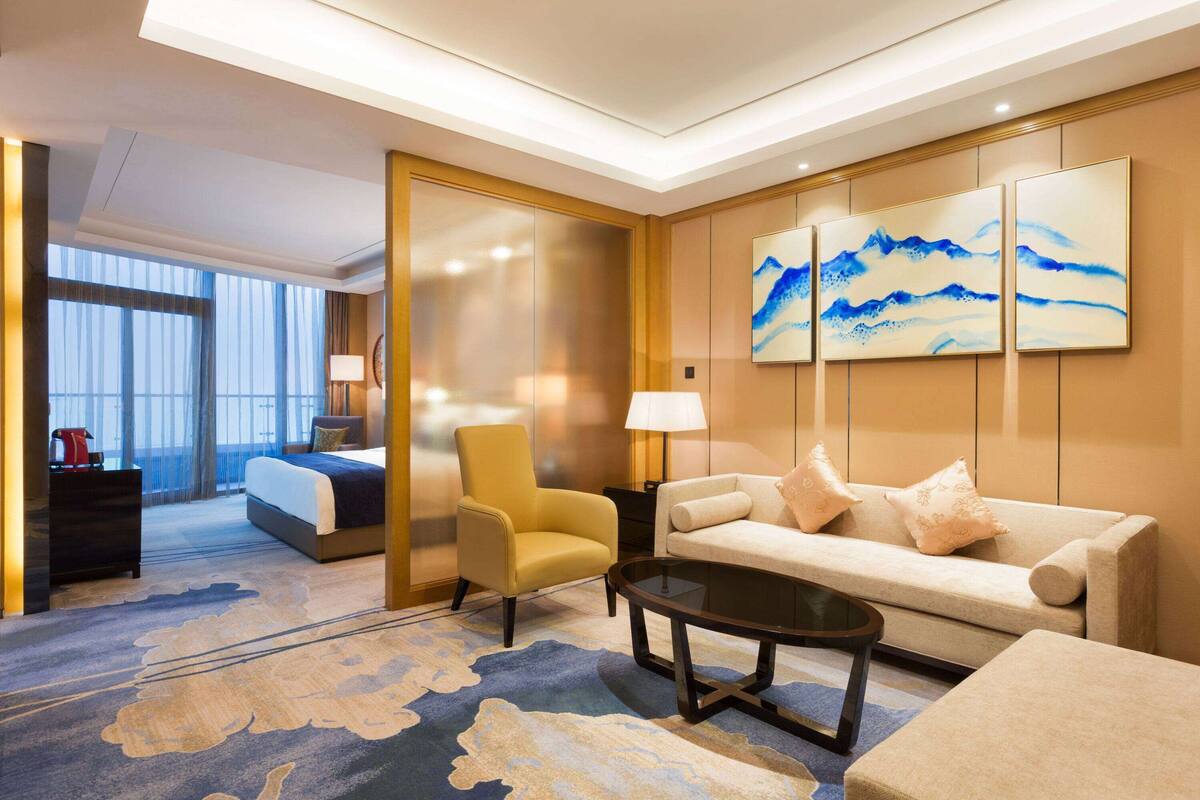 Un cuarto de hotel con una cama, un sillón amarillo, un sofá, una mesa de centro y obras de arte en la pared.