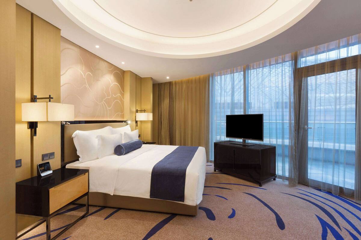 Habitación de hotel con una cama grande, mesitas de noche con lámparas, un televisor de pantalla plana y un ventanal con cortinas.