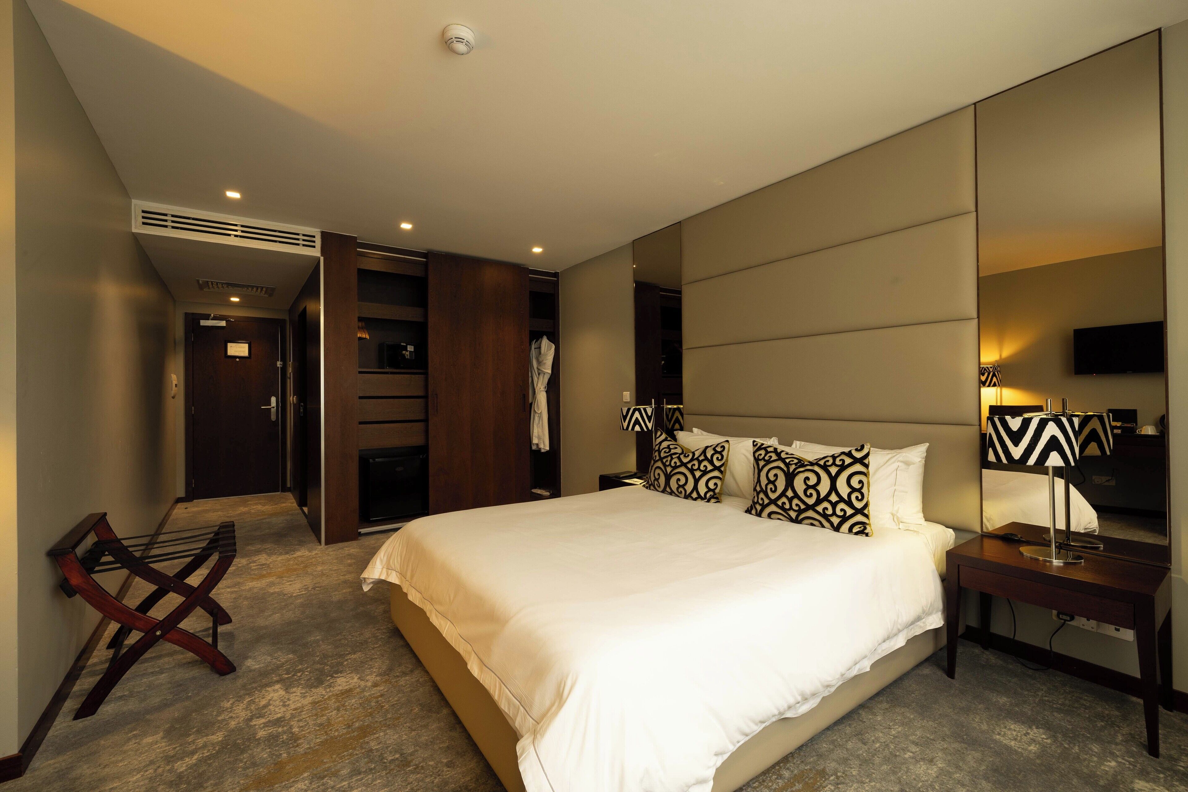 A modern hotel room with a large bed, a bedside table with a lamp, a wardrobe, and a mirror.