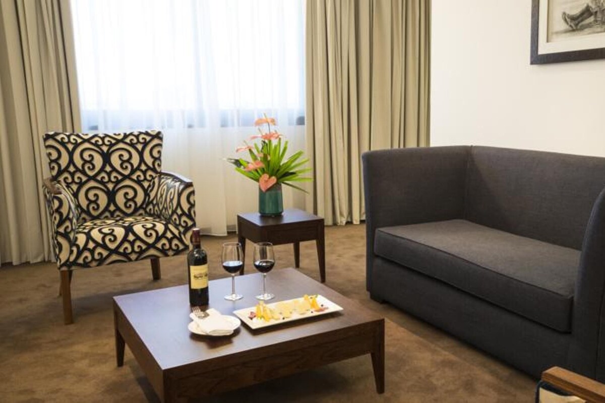 Una sala de estar con un sofá gris oscuro, un sillón estampado, una pequeña mesa de centro de madera con una botella y vasos, y un jarrón con flores.