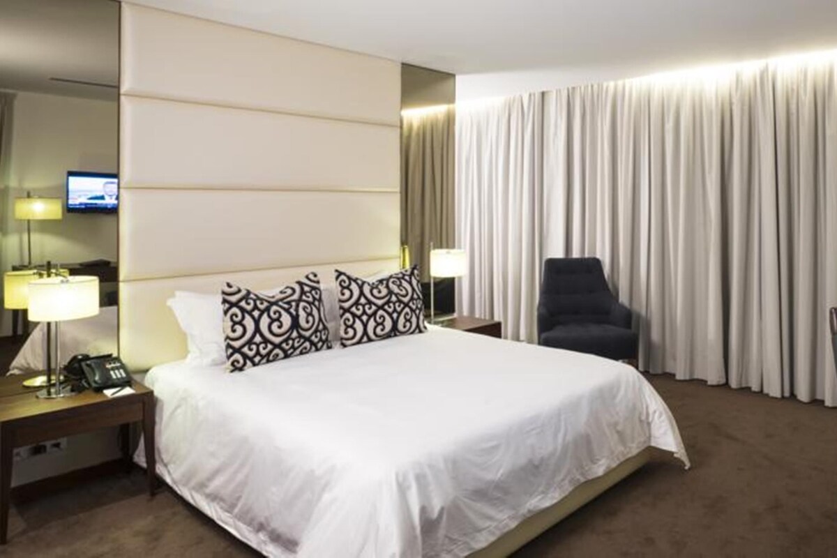 A hotel room with a large bed, two bedside lamps, a chair, a desk with a lamp, and a TV on the left wall.