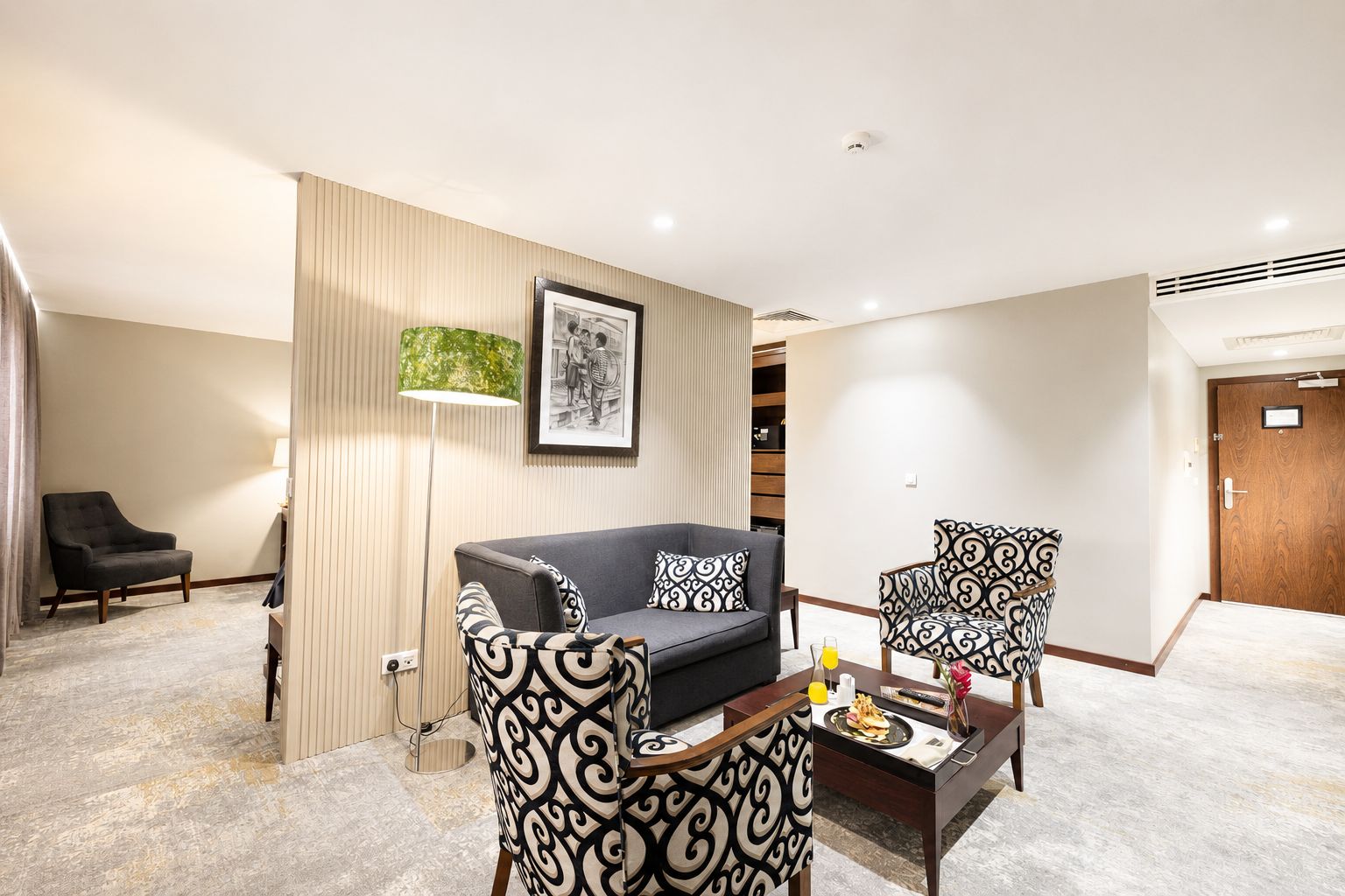 A hotel room with a sofa, two patterned armchairs, a coffee table, a lamp, and a framed picture on the wall.
