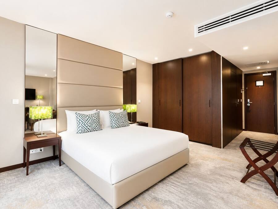 A modern hotel room with a large bed, a bedside table with lamps, a wardrobe, and a chair.
