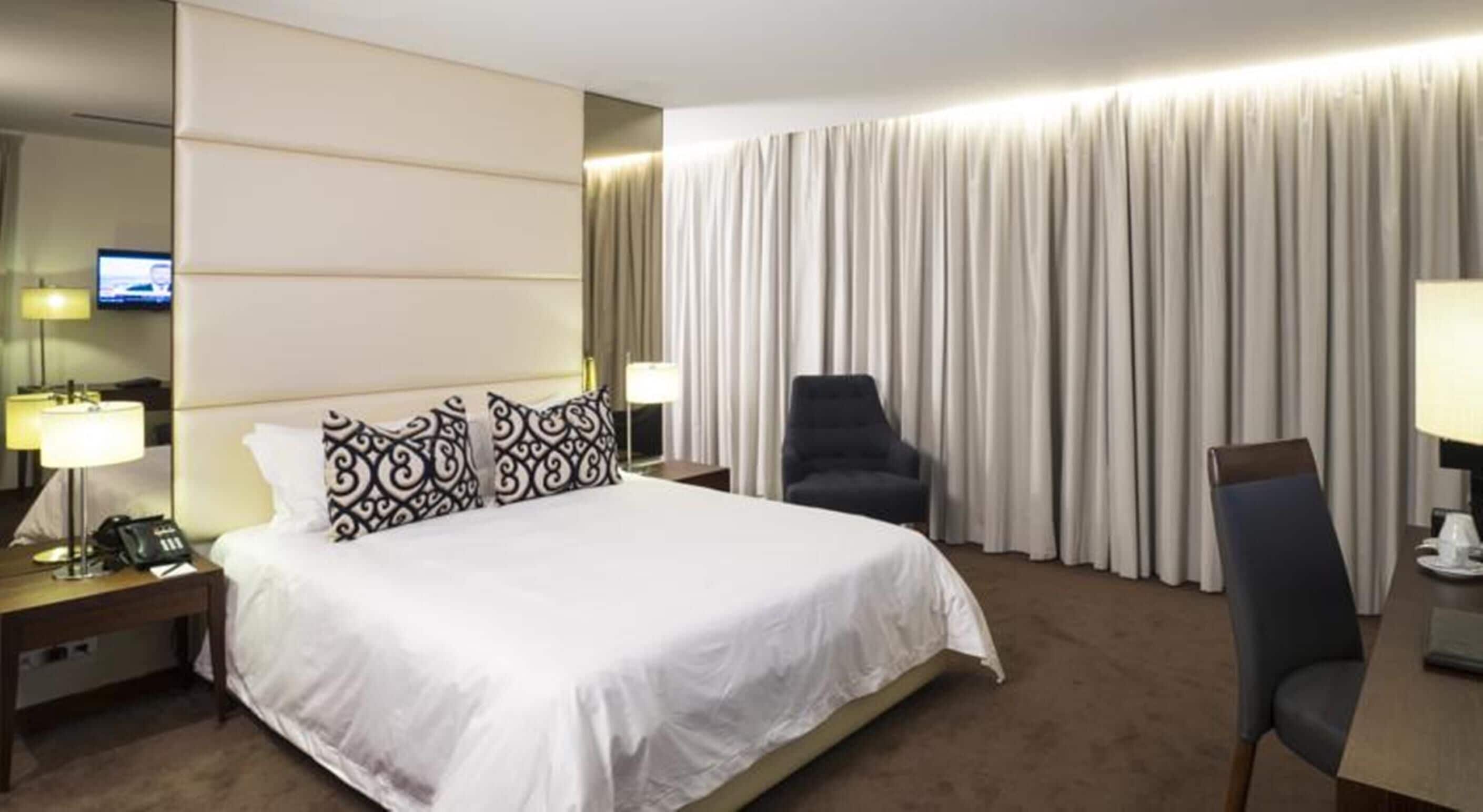 A hotel room with a large bed, a desk with a lamp, a chair, and a television on the wall.