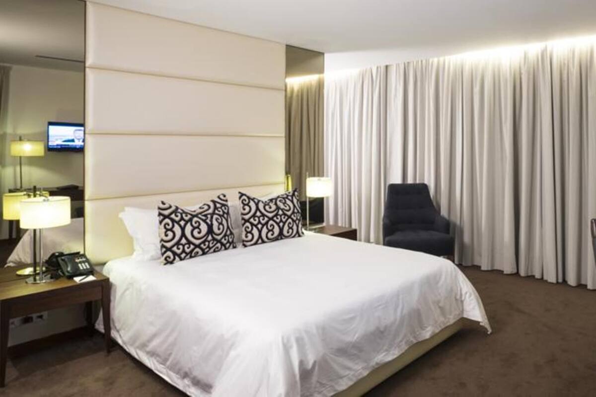 A hotel room with a large bed, two bedside lamps, a chair, a desk with a lamp, and a TV on the left wall.