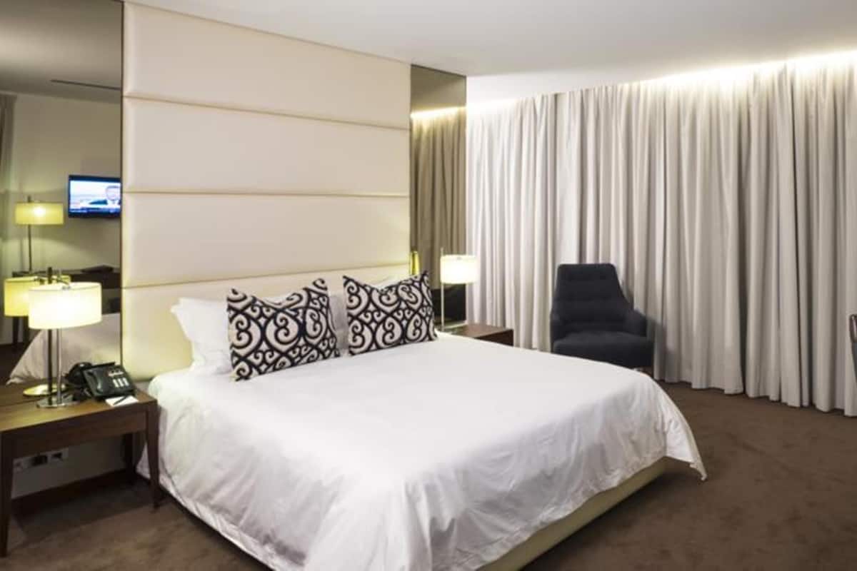 A hotel room with a large bed, a desk with a lamp, a chair, and a television on the wall.