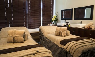 A hotel room with two beds, each with towels and a dark blanket. There is a bathroom with a sink, mirror, and toiletries.