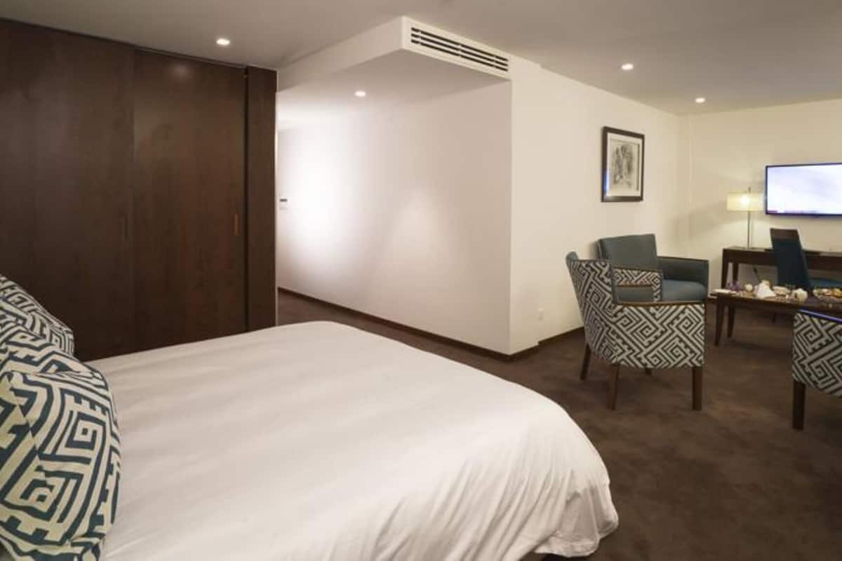 A hotel room with a large bed, a desk with a chair, a television, and a wall-mounted air vent.