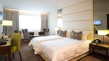 A hotel room with a large bed, two bedside tables, a desk with a chair, a TV, and a window with curtains.