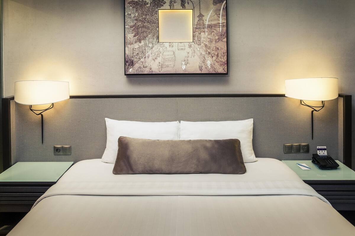 Habitación de hotel con una cama, dos lámparas de noche, una mesita con un teléfono y un cuadro en la pared.