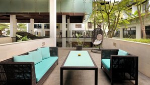 Un espacio exterior moderno con mobiliario de mimbre y una mesa central, rodeado de follaje y una estructura de edificio de varios niveles.