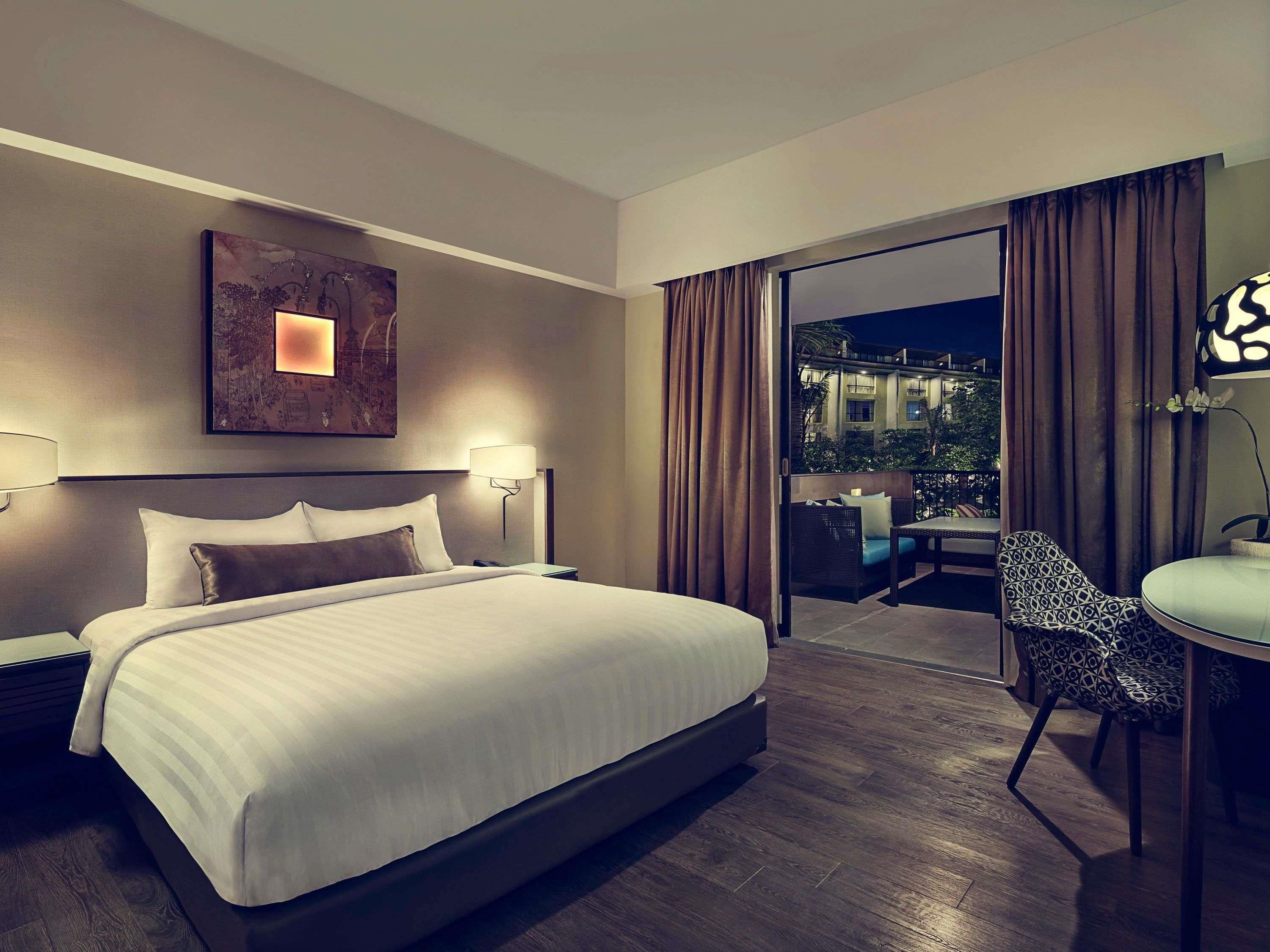 A hotel room with a large bed, a desk with a chair, a nightstand, a wall-mounted lamp, and a view of the outside.