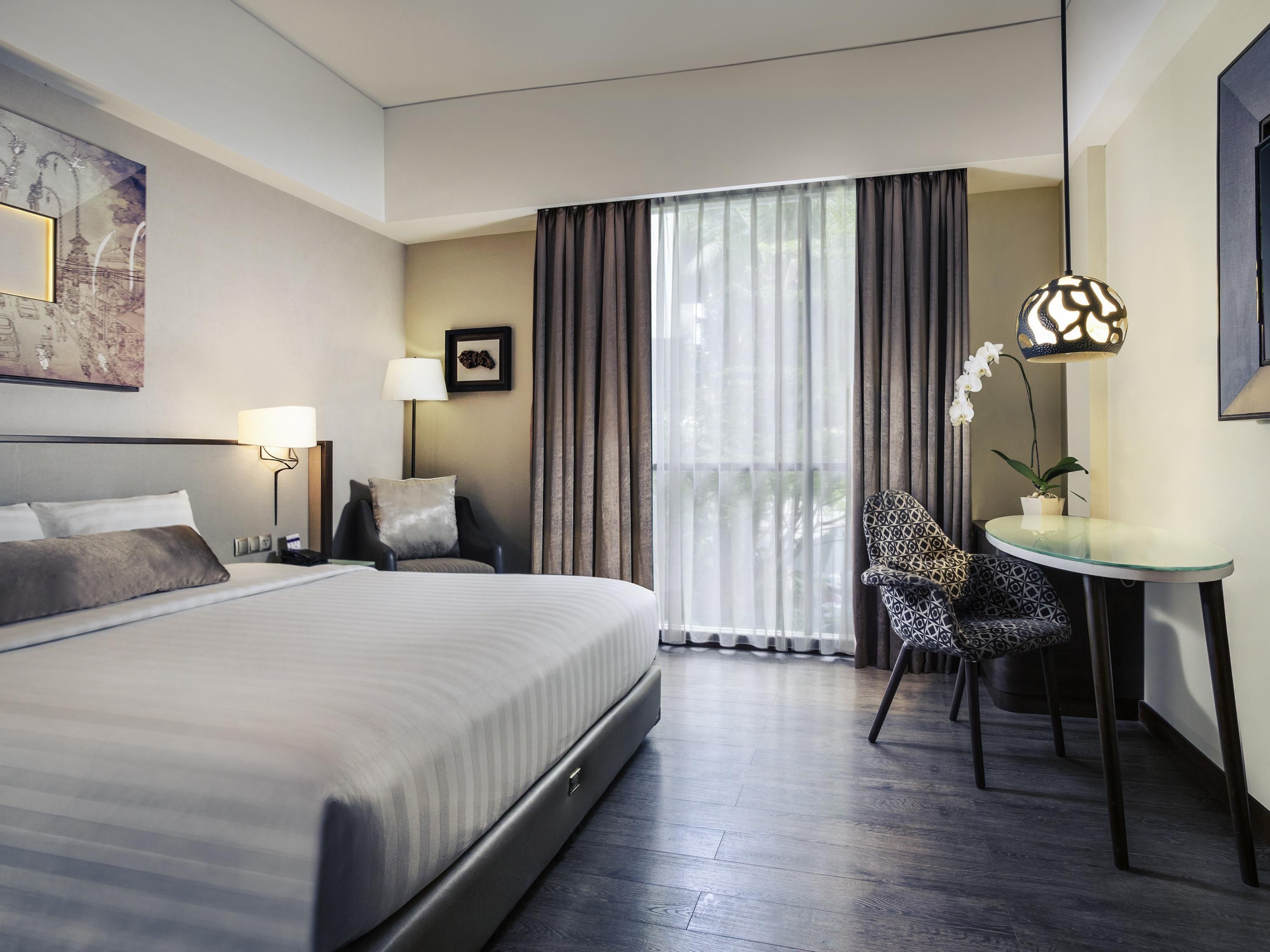 A hotel room with a large bed, a desk, a chair, a lamp, and a window with curtains.