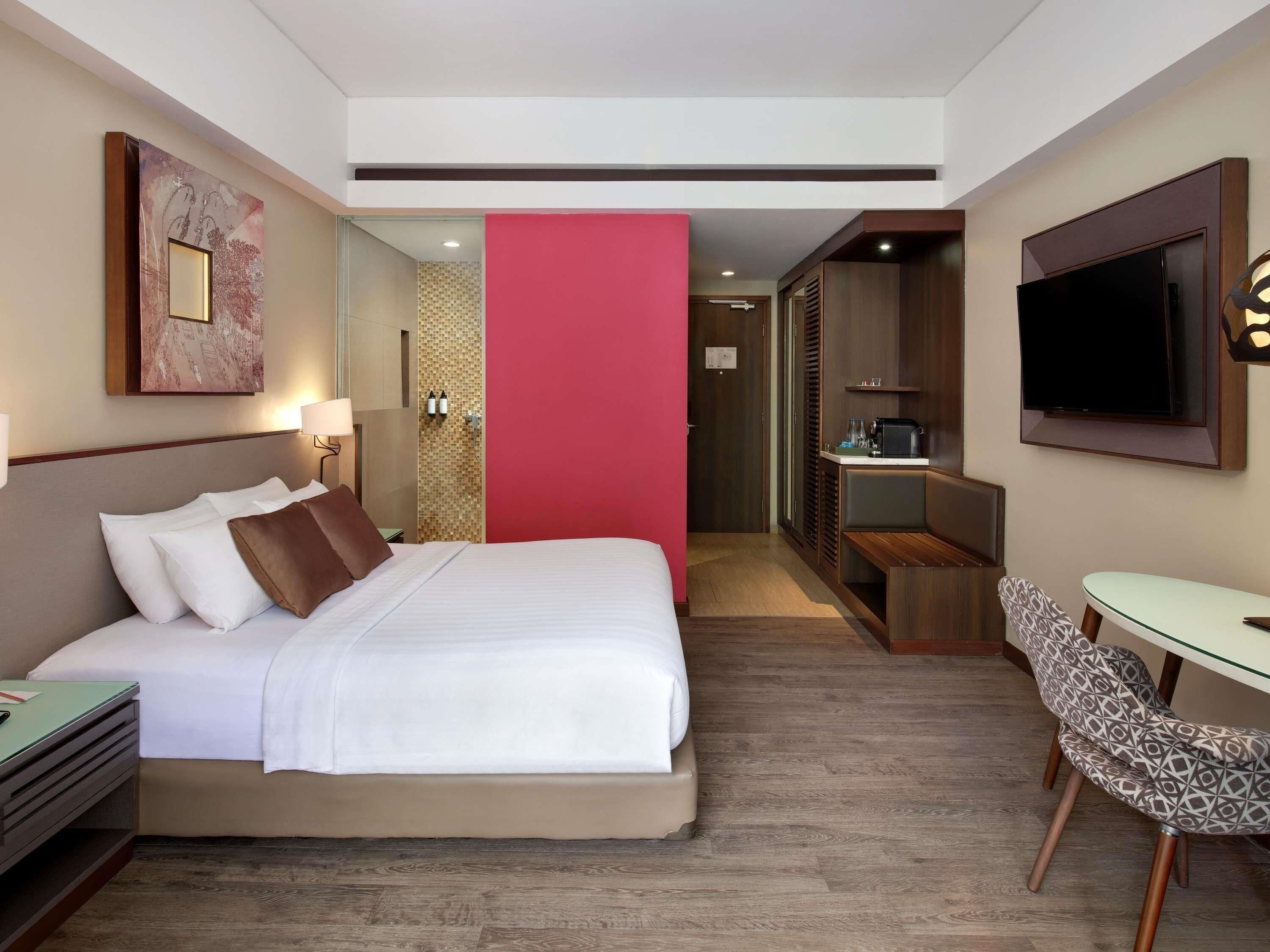 A hotel room with a bed, bedside table, lamp, TV, and a desk with a chair.