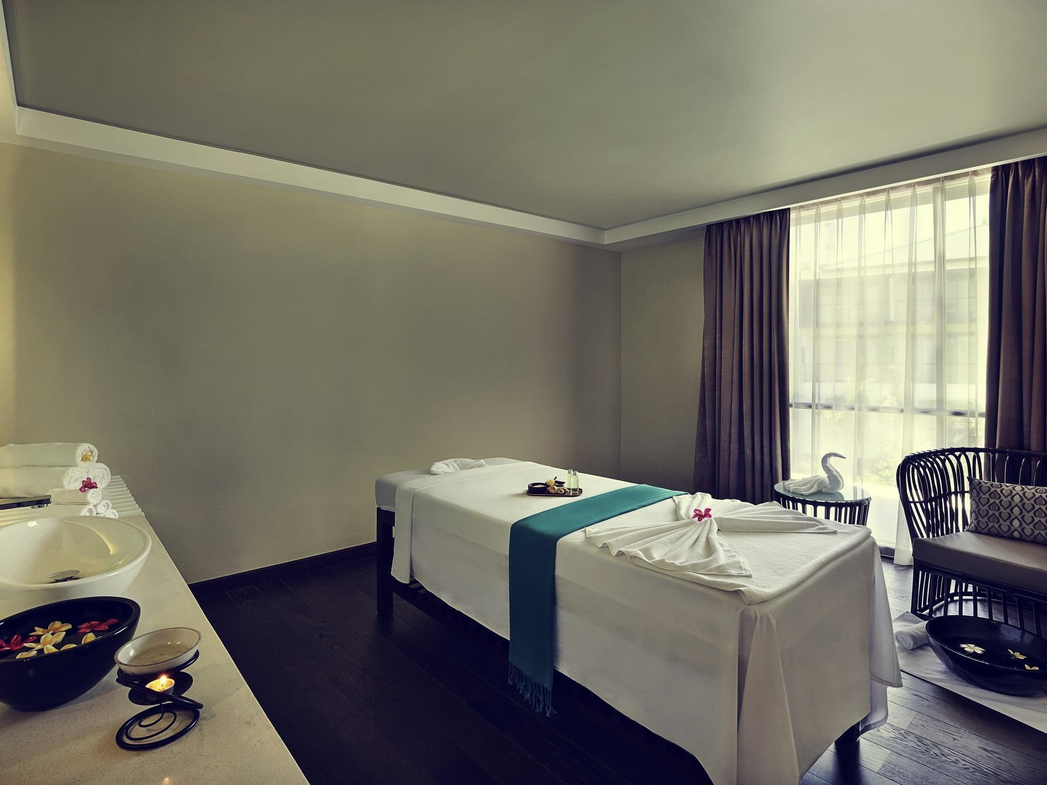 A hotel room with a bed, a small table with a bowl of flowers, a chair, and a window with curtains.