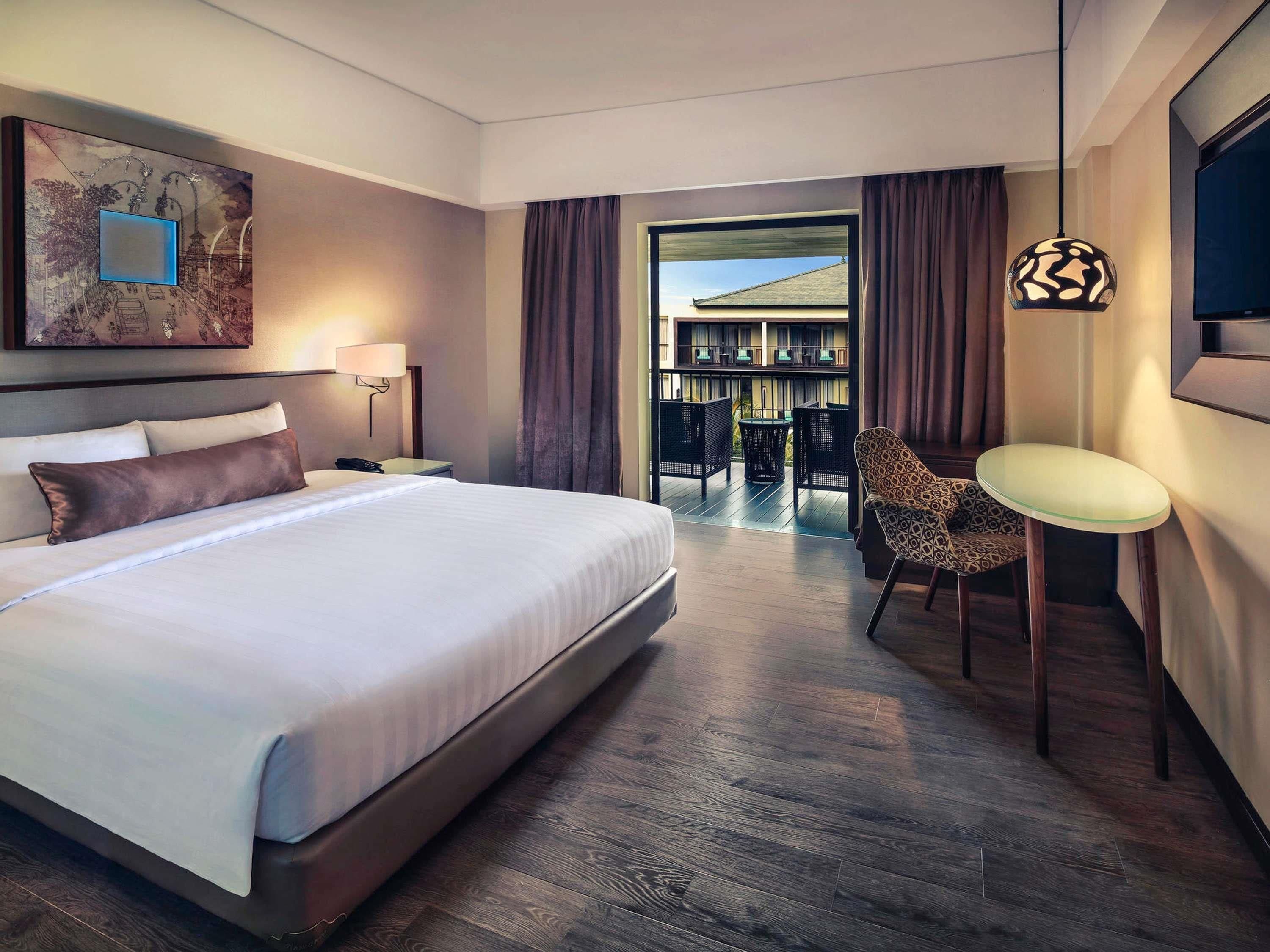 A hotel room with a large bed, a desk, a chair, a TV, and a balcony with a view.
