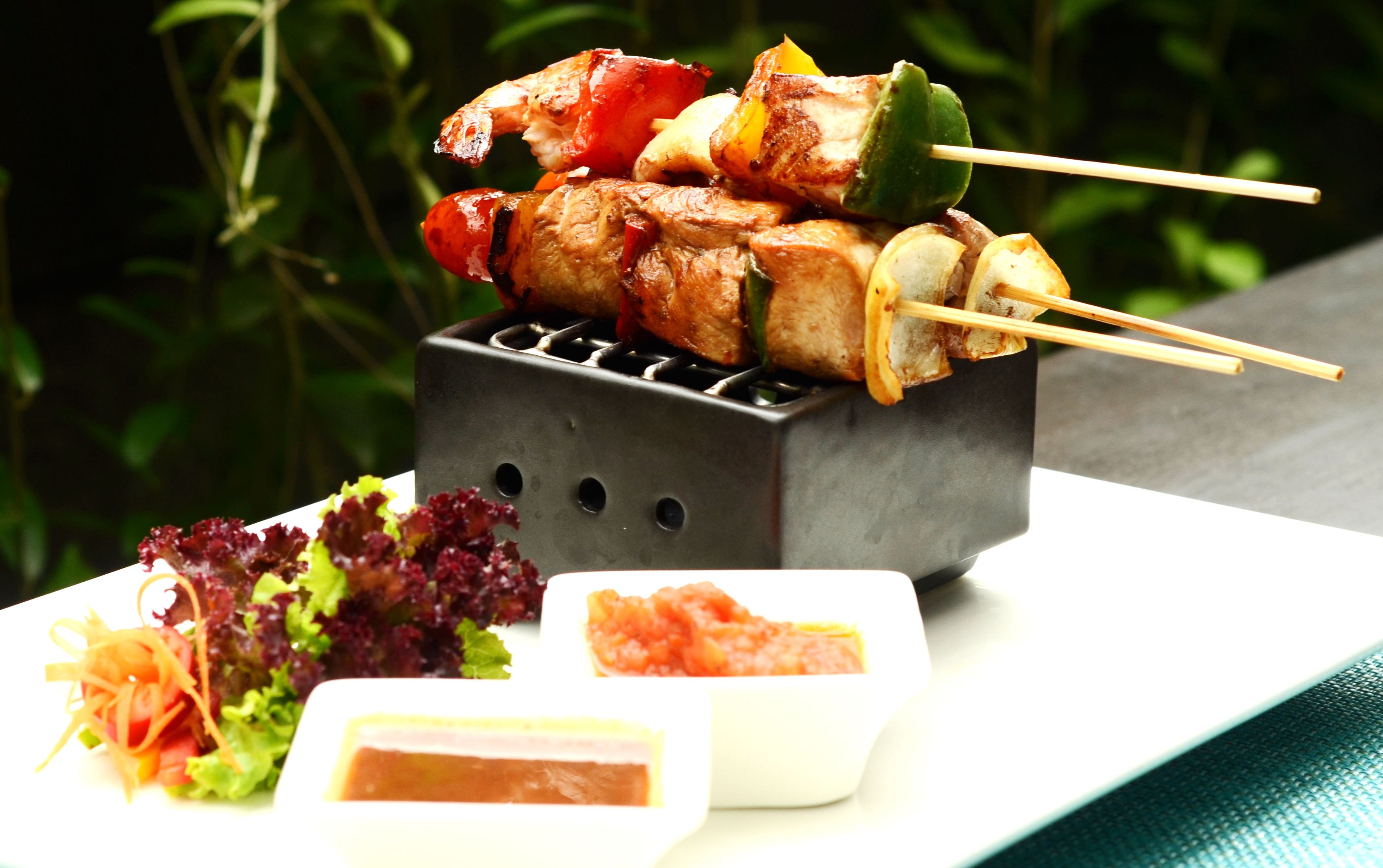 A grill with skewers of meat and vegetables, a plate of salad, and a small dish of sauce.