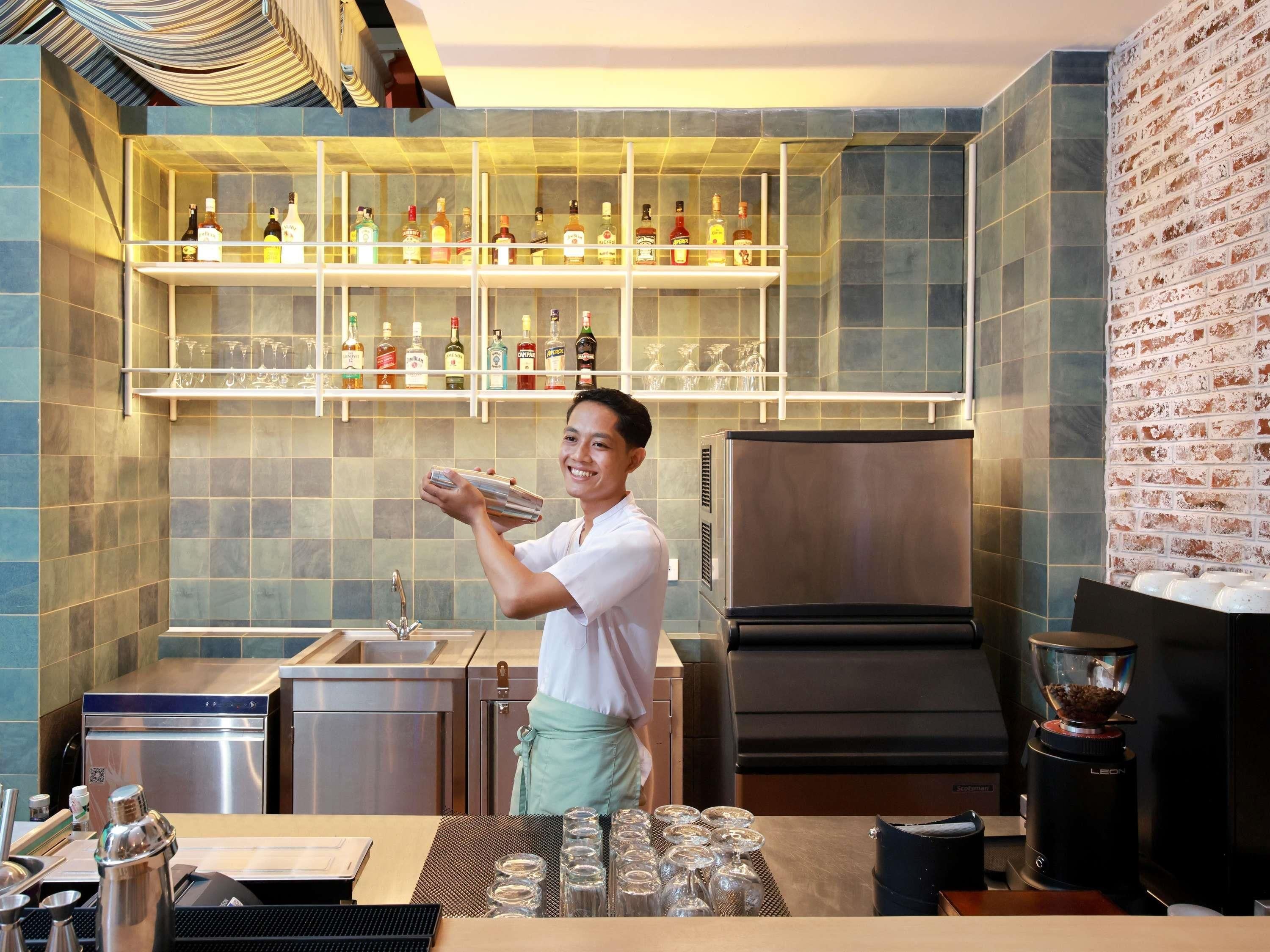 Una zona de bar con estantes llenos de botellas de licores, un barista preparando bebidas, una máquina de café y una pared de ladrillo.