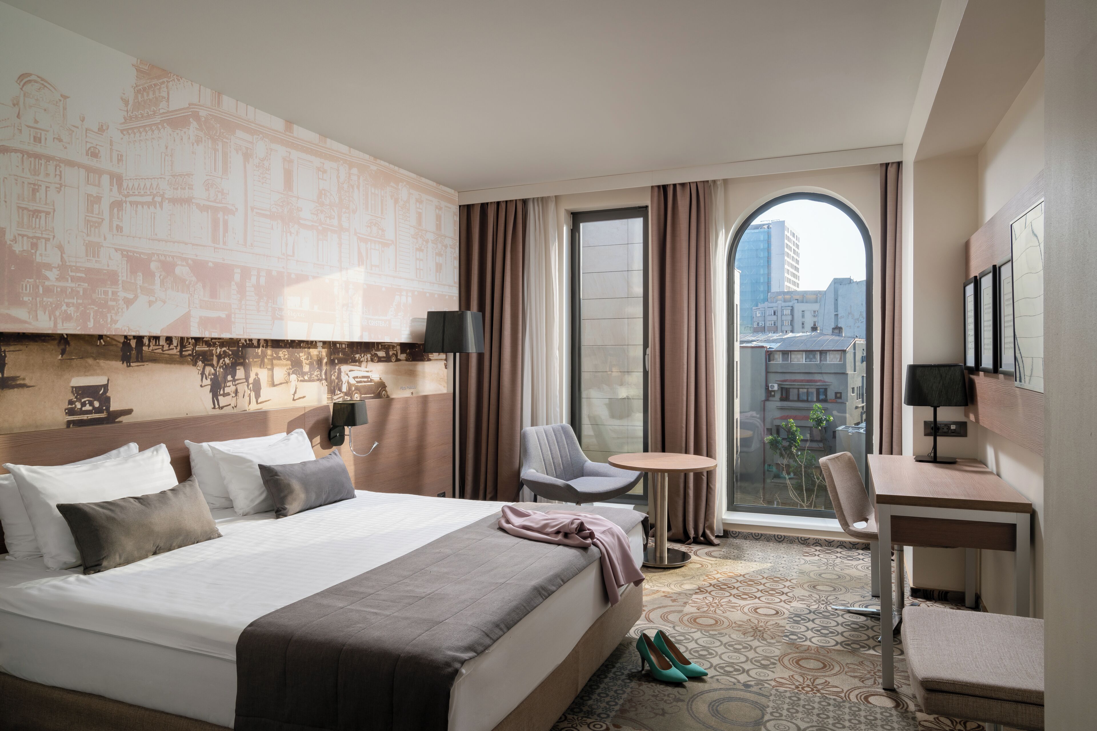 Habitación de hotel con una cama grande, un escritorio, una silla y vistas de la ciudad a través de un ventanal amplio.