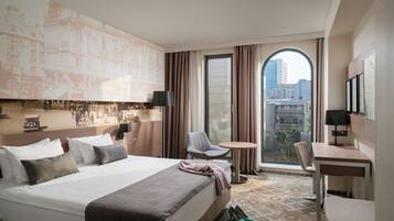 Habitación de hotel con una cama grande, un escritorio, una silla y vistas de la ciudad a través de un ventanal amplio.
