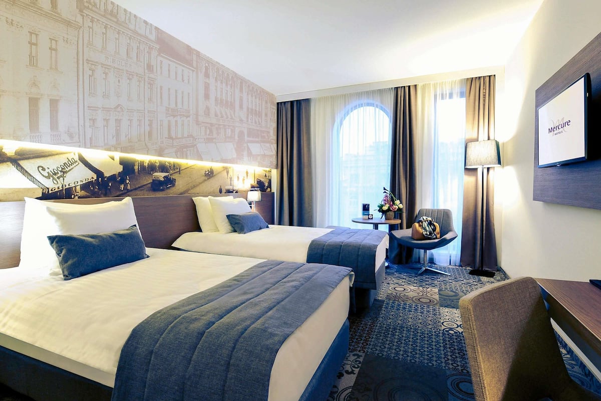 A hotel room with two beds, a desk, a chair, a lamp, and a large window with curtains.