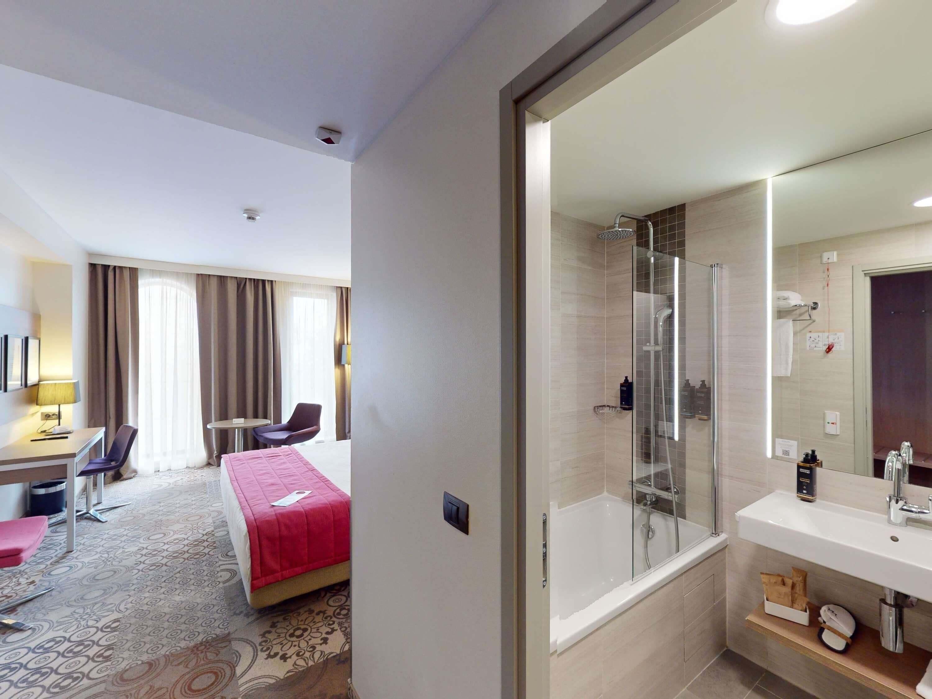 A hotel room with a bed, a desk, a chair, a shower area, and a bathroom with a sink and mirror.