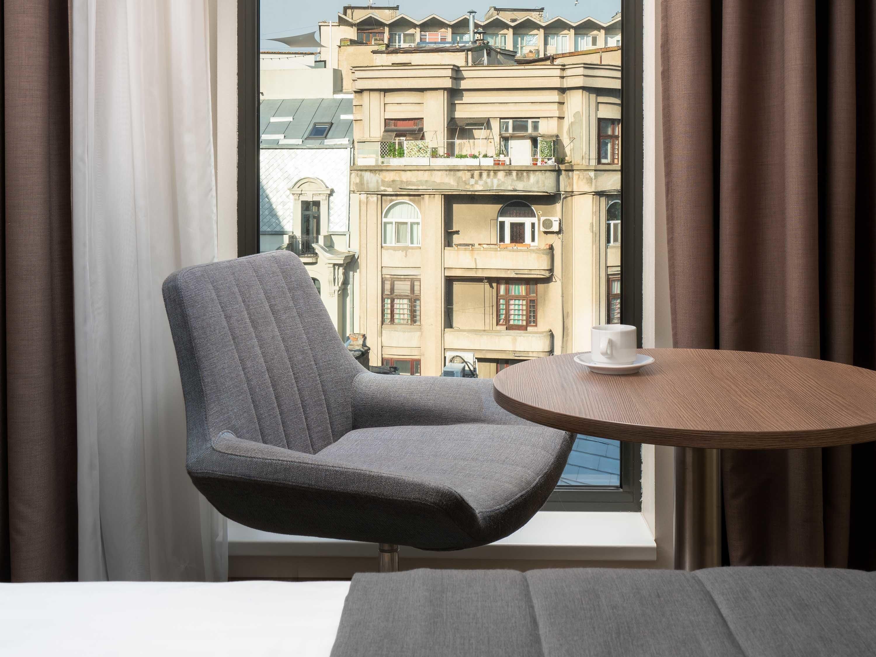 A modern room with a grey upholstered chair, a round wooden table with a white cup, and a view of a building through a window with brown curtains.