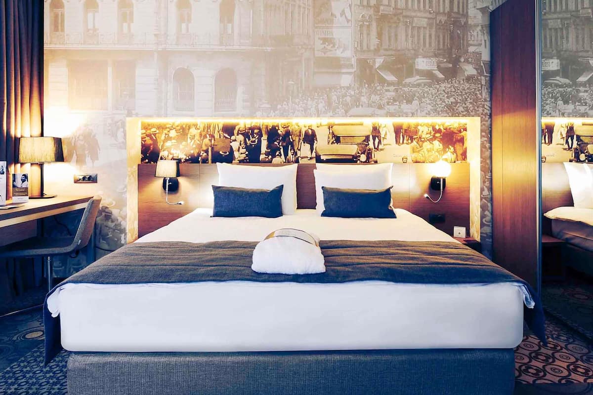 A hotel room with a large bed, two bedside tables, a desk, and a chair. The room features a large mural of a historical scene on the wall behind the bed.