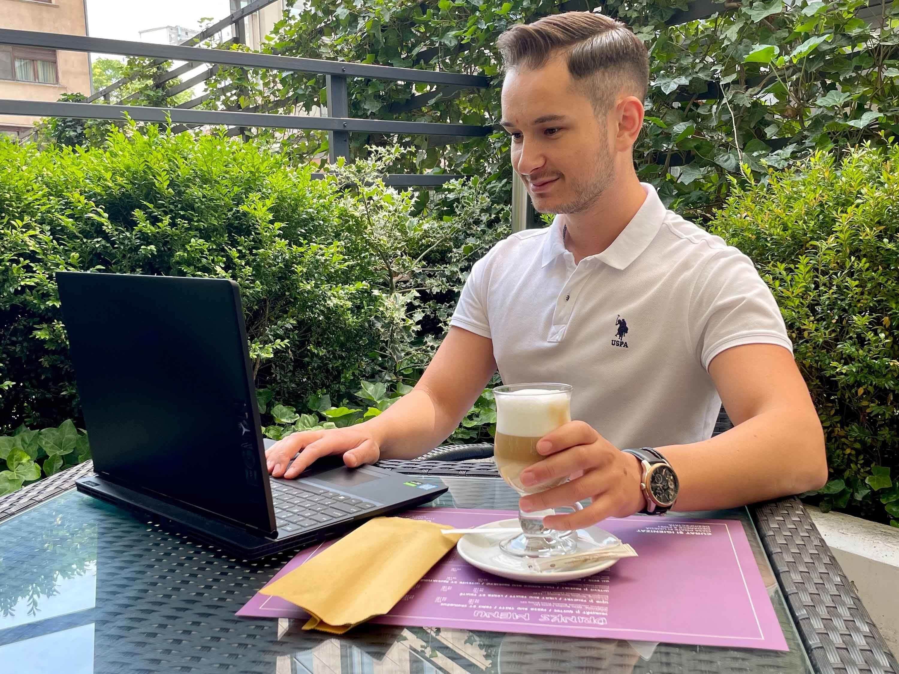 A man sitting at an outdoor table with a laptop, holding a glass of beer, and wearing a white polo shirt with a logo.