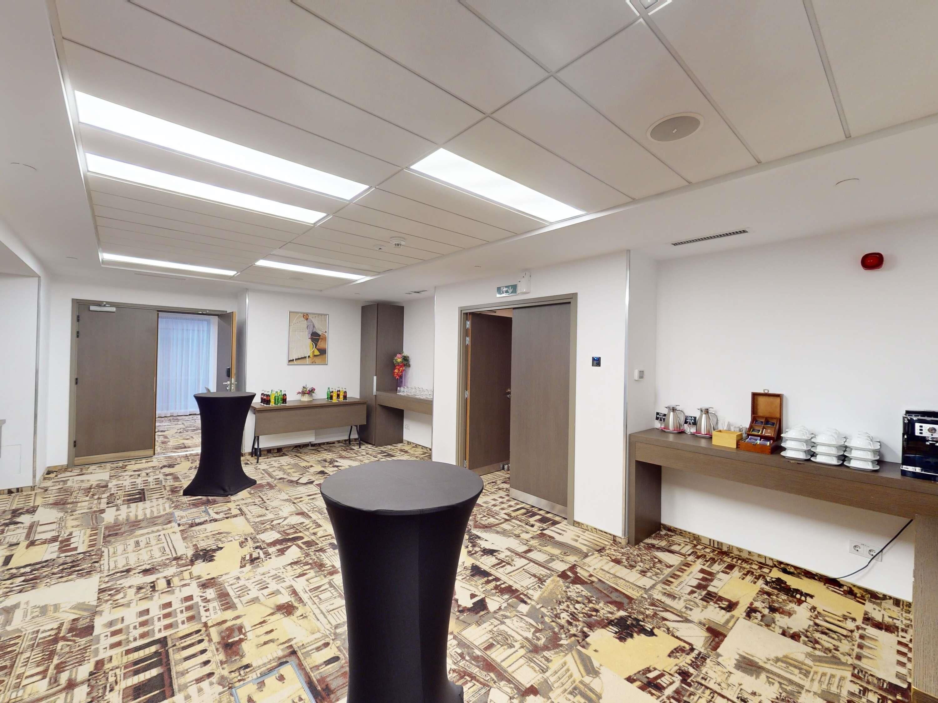 A hotel room with a patterned carpet, a black round table, a long table with bottles, and a wall-mounted shelf with items.