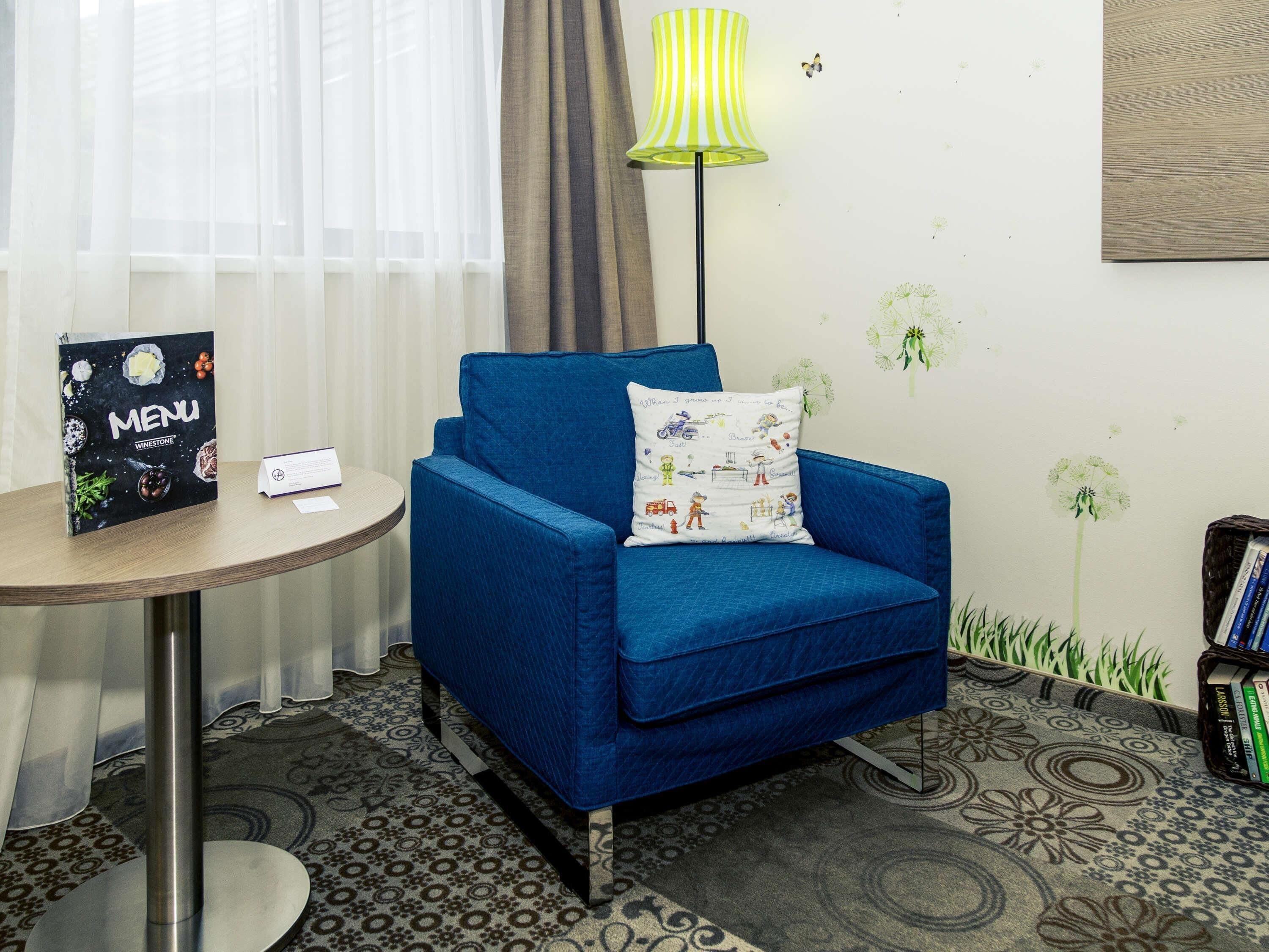Un fauteuil bleu avec un coussin blanc, une table ronde avec un menu, une lampe et une bibliothèque.
