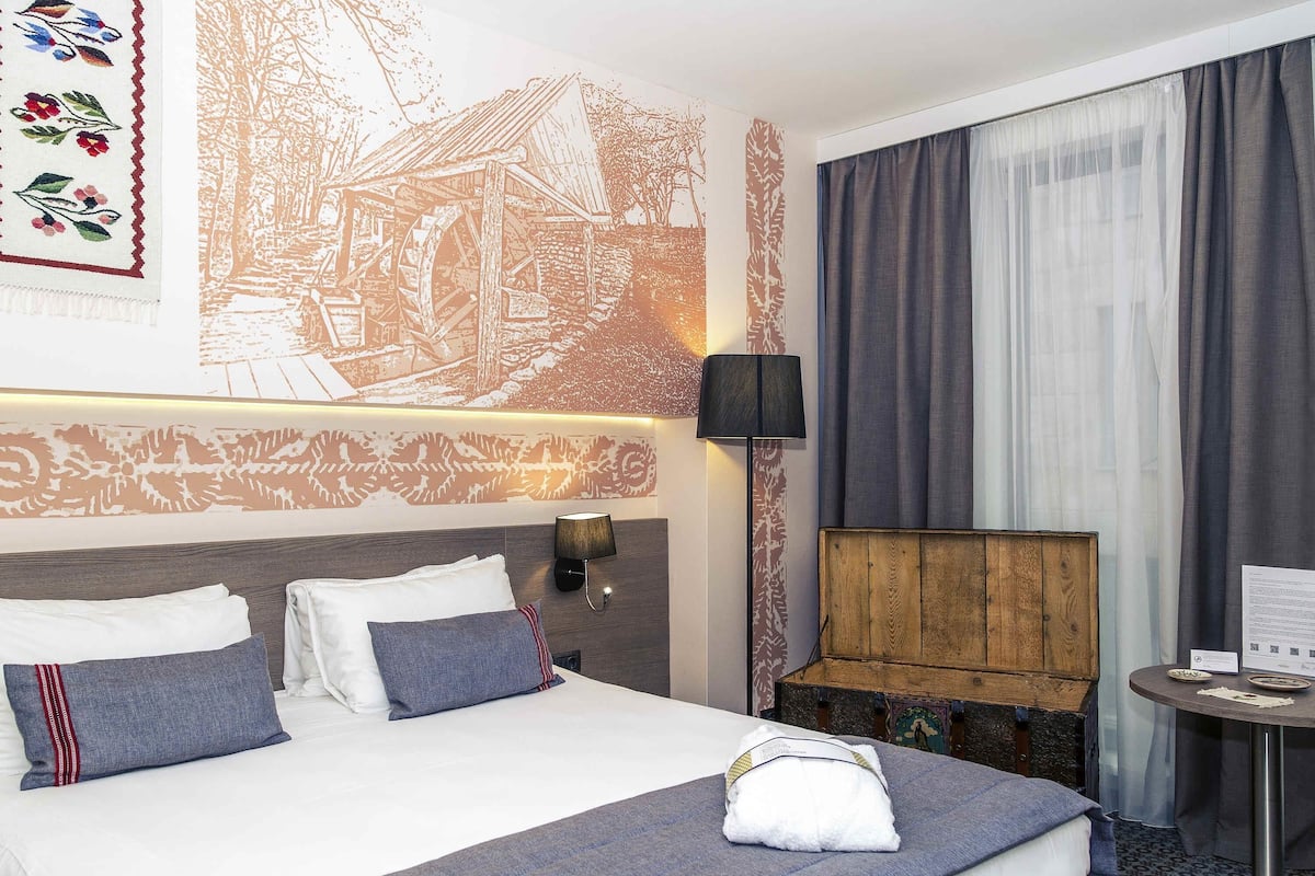 A hotel room with a large bed, a wooden headboard, a wall-mounted lamp, a small table with a menu, and a window with curtains.