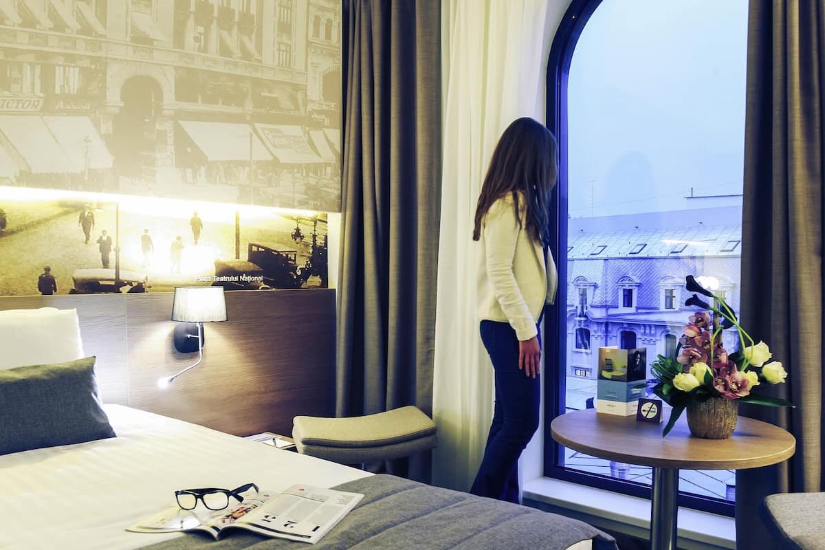 A hotel room with a bed, a small table with a vase of flowers, a lamp, and a view of buildings through a large window.