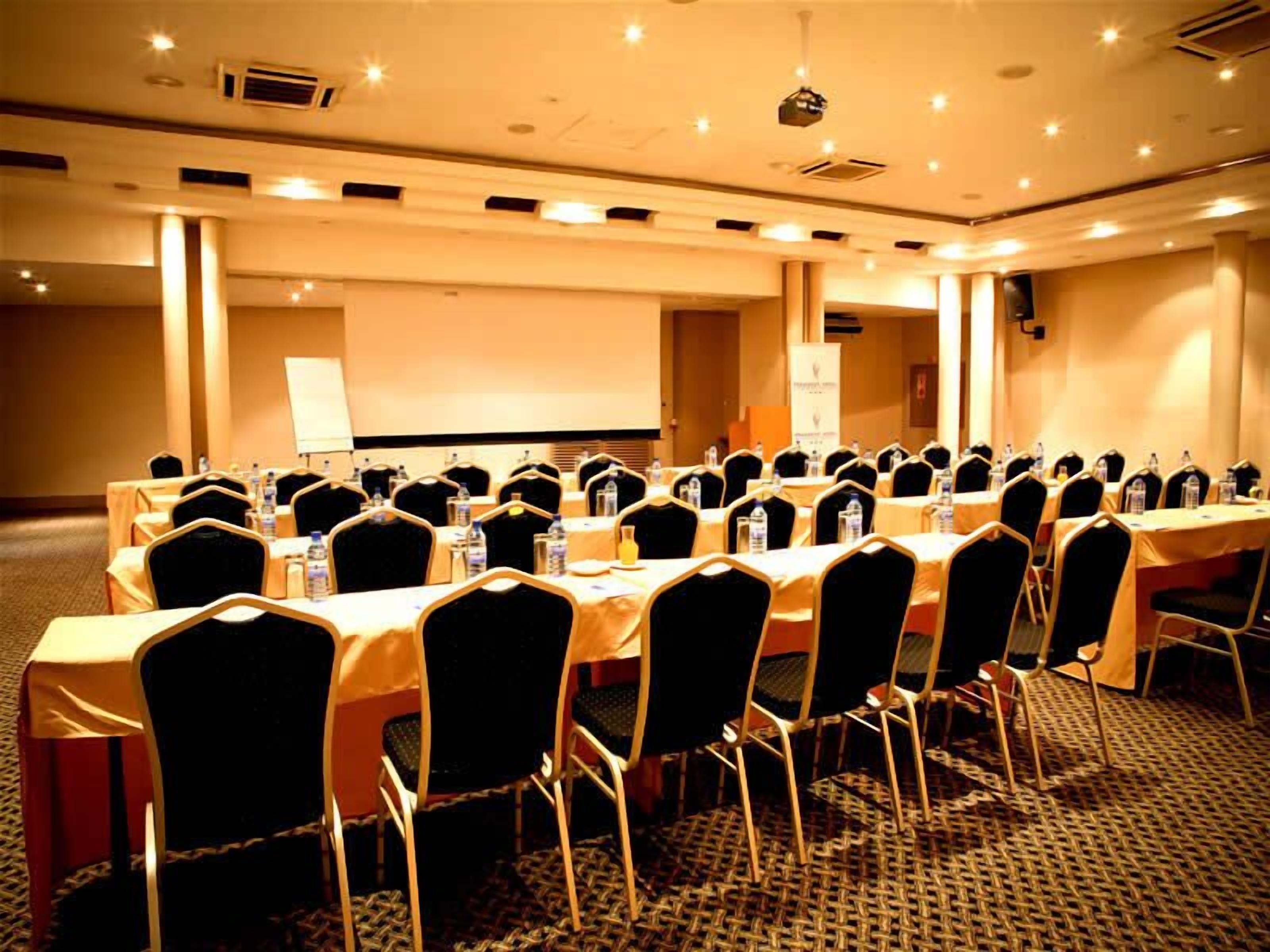 A conference room with rows of tables and chairs, equipped with water bottles and a projector screen.