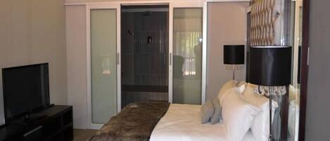 Ein modernes Hotelzimmer mit einem großen Bett, einem Fernseher, einer Kristallleuchtre und einer Schiebetür zu einem Duschbereich.