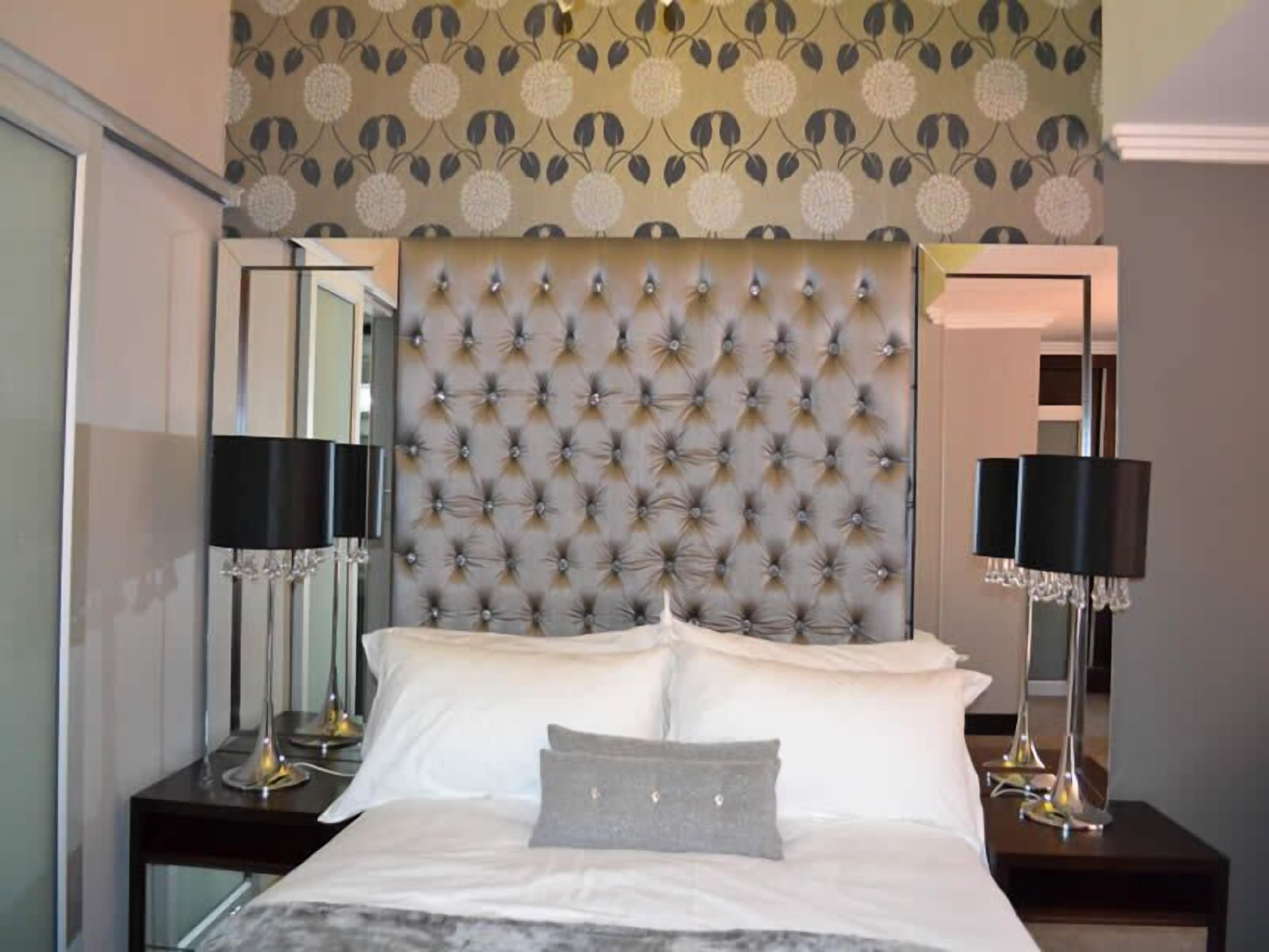 Una habitación de hotel con cabecera capitonada, un espejo grande, dos mesitas de noche con lámparas y una cama con sábanas blancas y almohada gris.