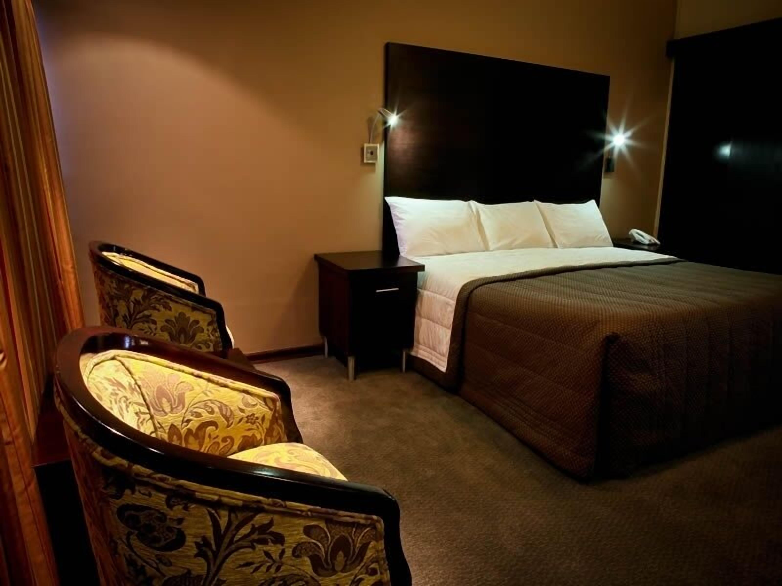 A hotel room with a bed, nightstand, lamp, and two upholstered chairs.