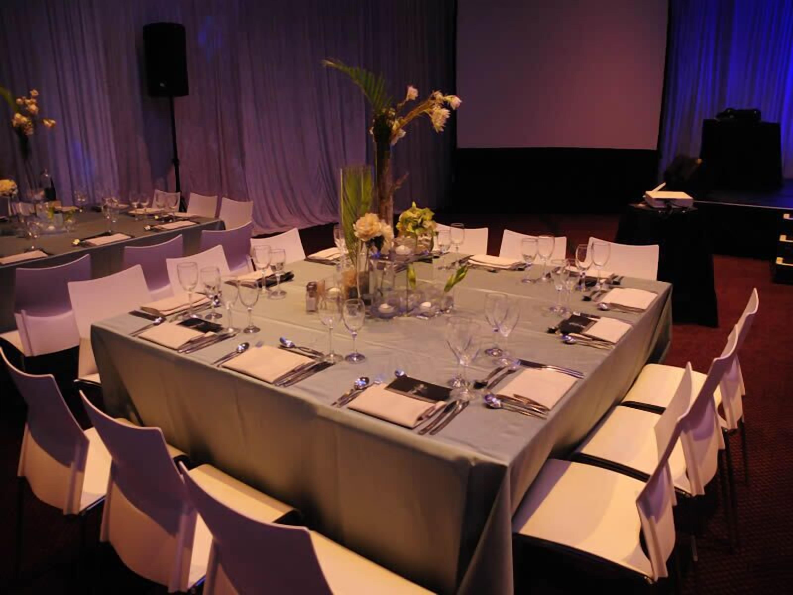 Un salón de banquetes con largas mesas rectangulares dispuestas para recibir comensales, cada una con sillas blancas y un arreglo floral central.
