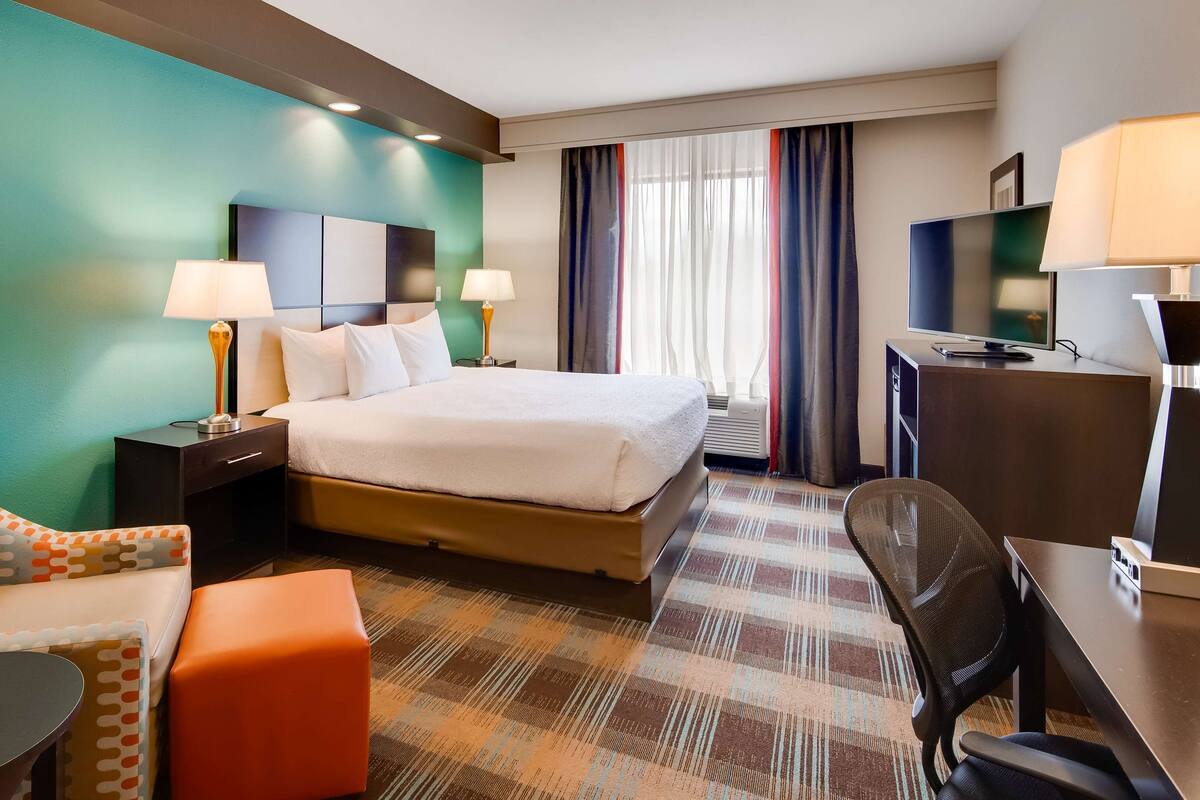 A hotel room with a bed, two bedside tables with lamps, a desk with a chair, a television, and a large window with curtains.