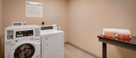 Zona de lavandería para huéspedes con lavadora y secadora de carga frontal, un cartel con instrucciones para la ropa y una mesa con detergente y toallas.