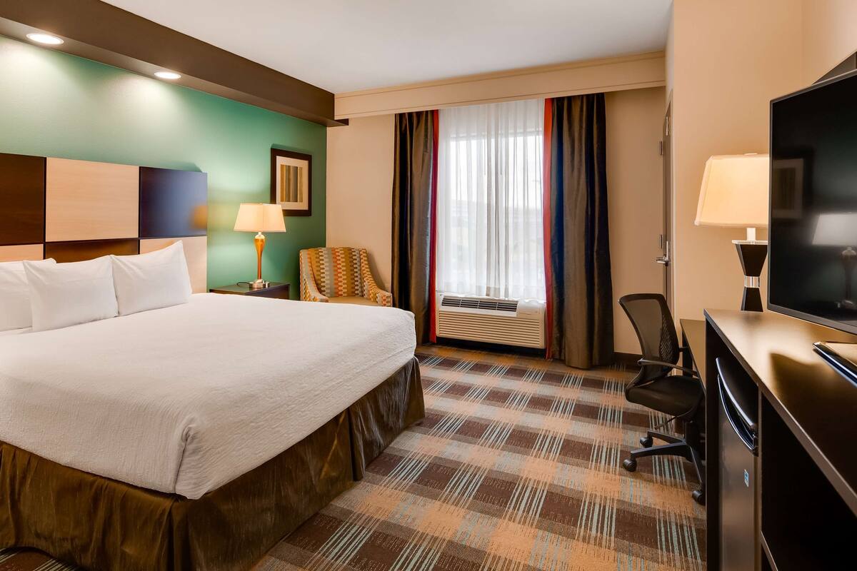 A hotel room with a large bed, a desk with a television, a chair, a lamp, and an air conditioning unit.