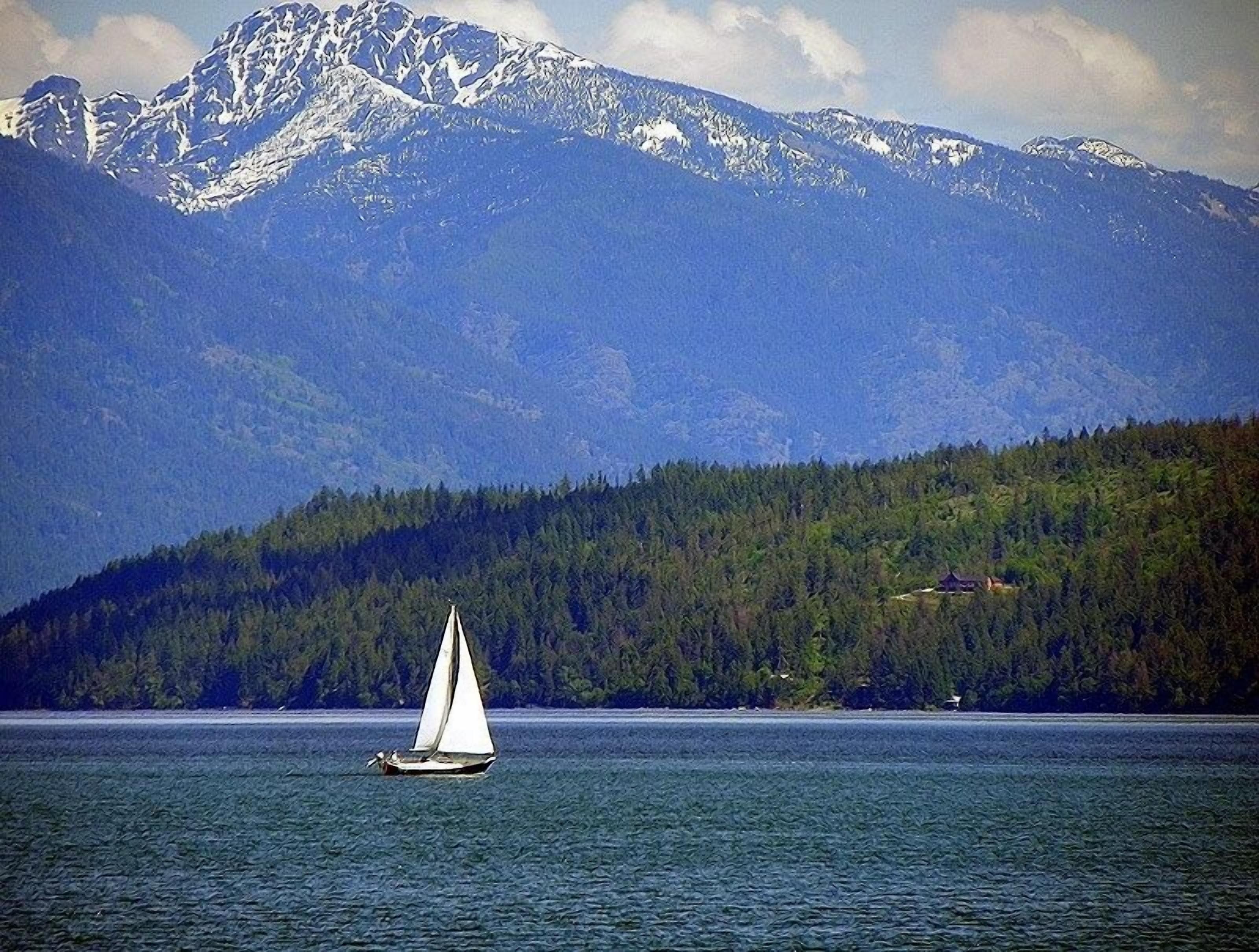 Un velero en un lago tranquilo, con una cadena montañosa boscosa al fondo.